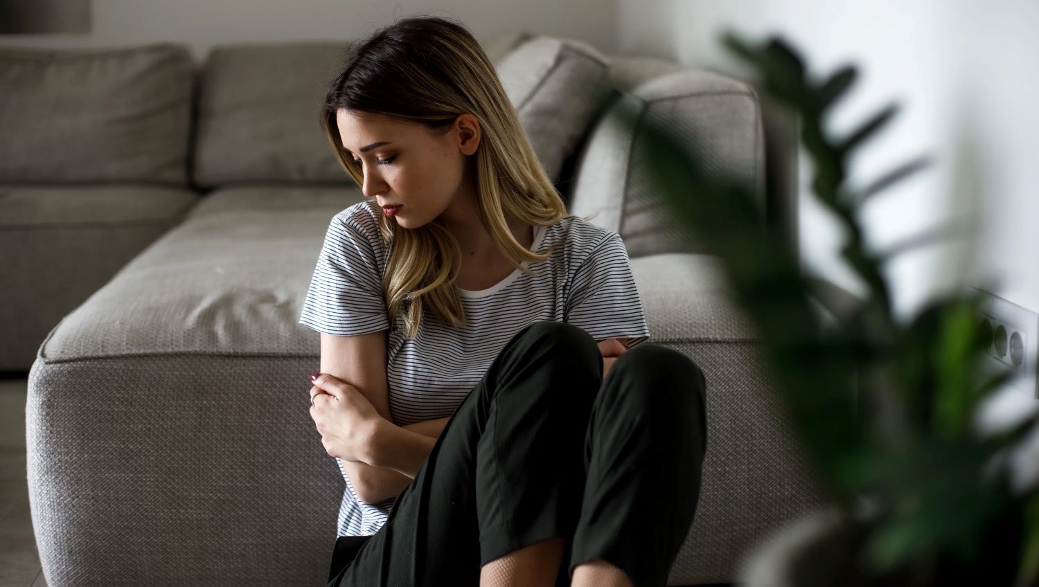 Donna triste seduta a terra, appoggiata al divano.