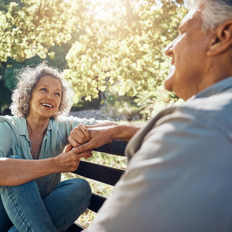 Un couple âgé souriant est assis sur un banc de parc sous le soleil. La femme aux cheveux gris bouclés tient la main de l'homme et le regarde. La scène dégage une atmosphère de calme et d'affection.